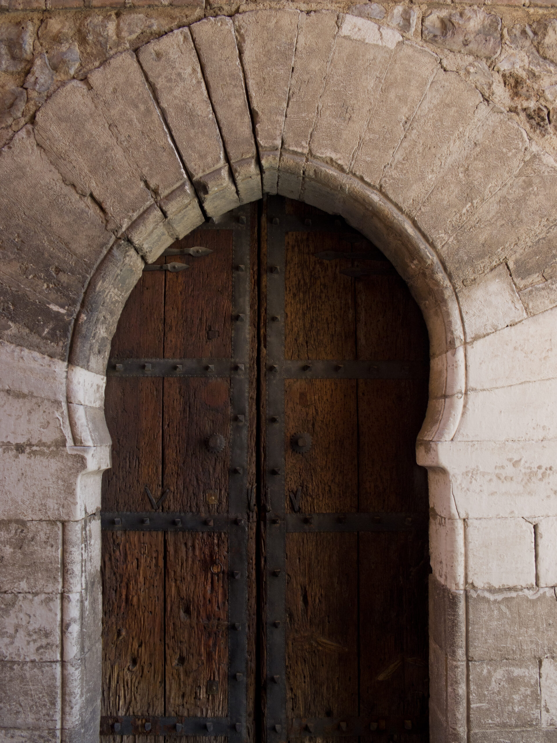 Puerta histórica de madera maciza con herrajes metálicos y clavos decorativos, enmarcada por un arco de piedra de sillería con dovelas, integrada en un muro de mampostería y piedra clara de estilo arquitectónico tradicional.