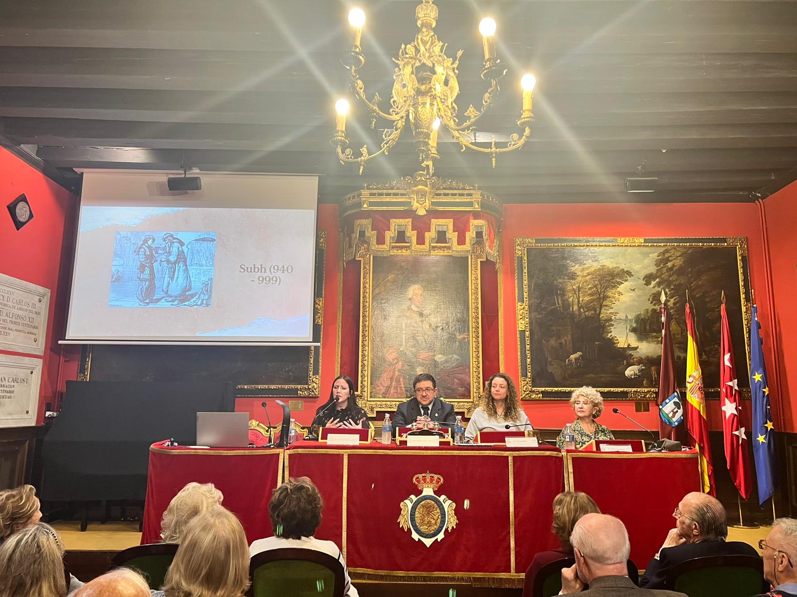 Fotografía horizontal frontal de ponentes y público en una presentación literaria con proyección en pantalla.