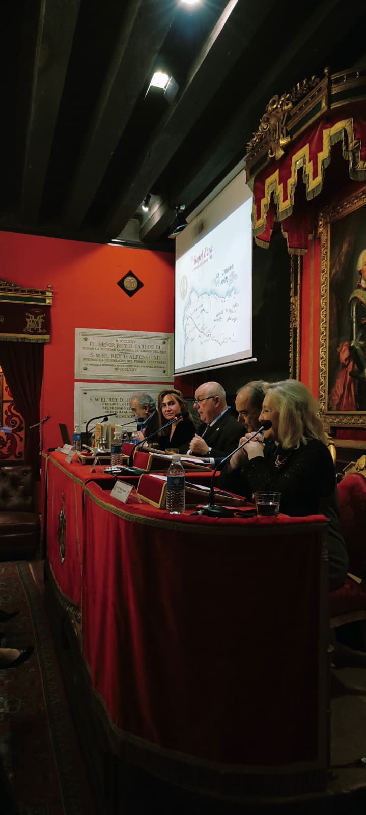 Fotografía vertical de mesa de ponentes durante una presentación literaria en salón histórico con pantalla al fondo.
