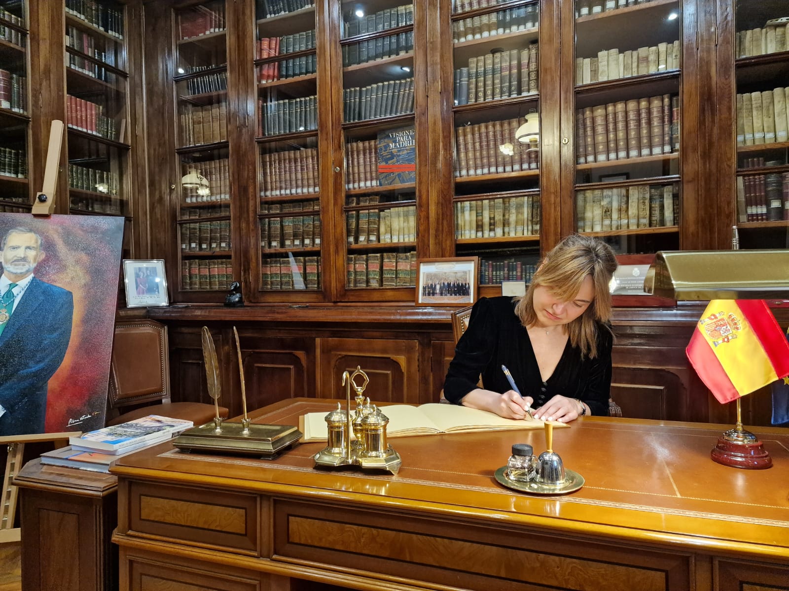 Fotografía horizontal de firma institucional en biblioteca histórica con retrato y mesa de despacho en primer término.