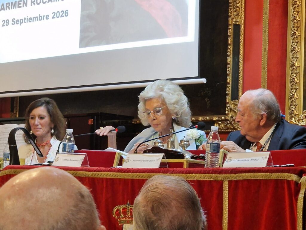 Fotografia de tres participantes sentados tras la mesa del acto Cuadros para una exposicion: Mussorgsky, con microfonos, documentacion y una proyeccion del evento al fondo.