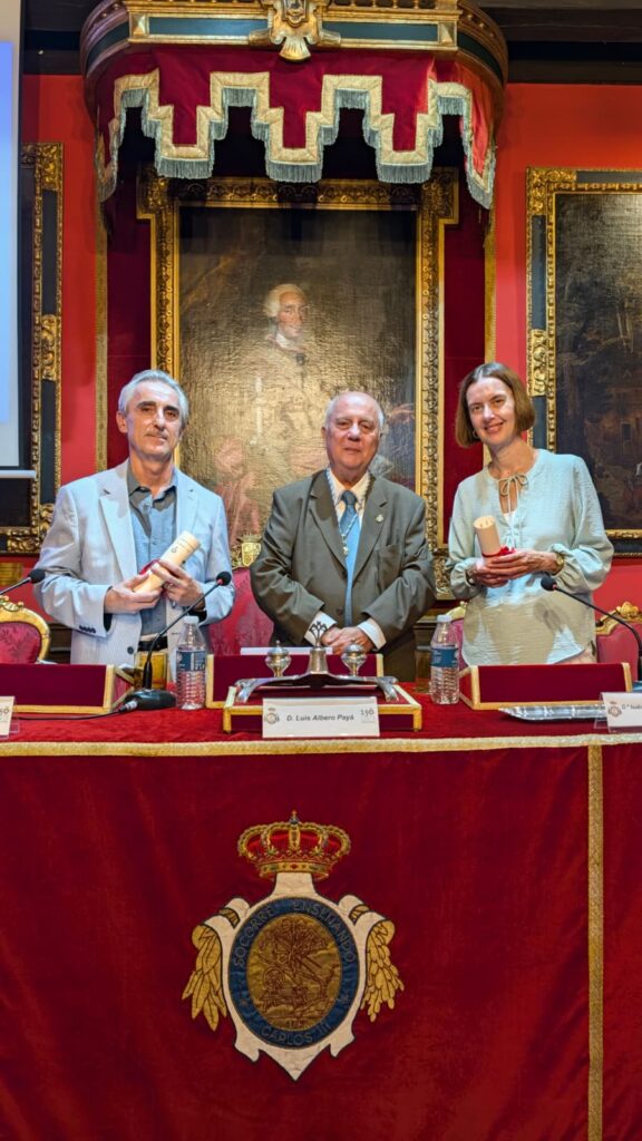 Fotografia vertical de tres participantes posando tras la mesa principal de un salon historico, con retratos enmarcados y decoracion institucional, durante el acto Una infanta fren