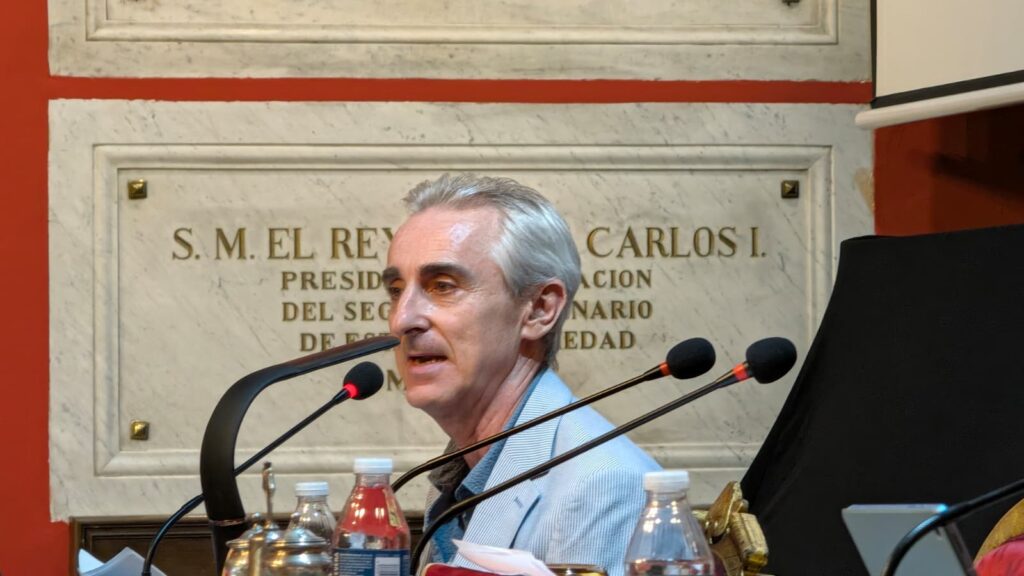 Primer plano de un ponente interviniendo ante varios microfonos durante el acto Una infanta frente a Napoleon, con una placa conmemorativa de la institucion al fondo.