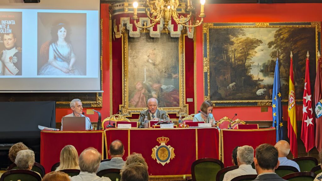 Vista general del acto Una infanta frente a Napoleon con mesa, publico y proyeccion. Fotografia general del salon durante el acto Una infanta frente a Napoleon. Tres participantes