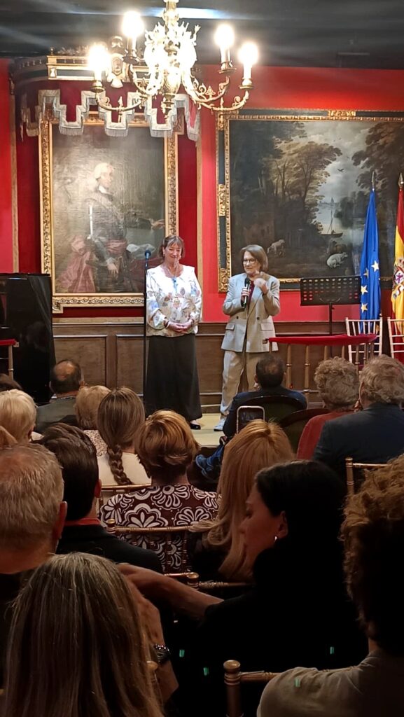Dos mujeres intervienen en el escenario del sal?n hist?rico de la Real Sociedad Econ?mica Matritense, con p?blico sentado frente a la mesa y decoraci?n institucional al fondo.