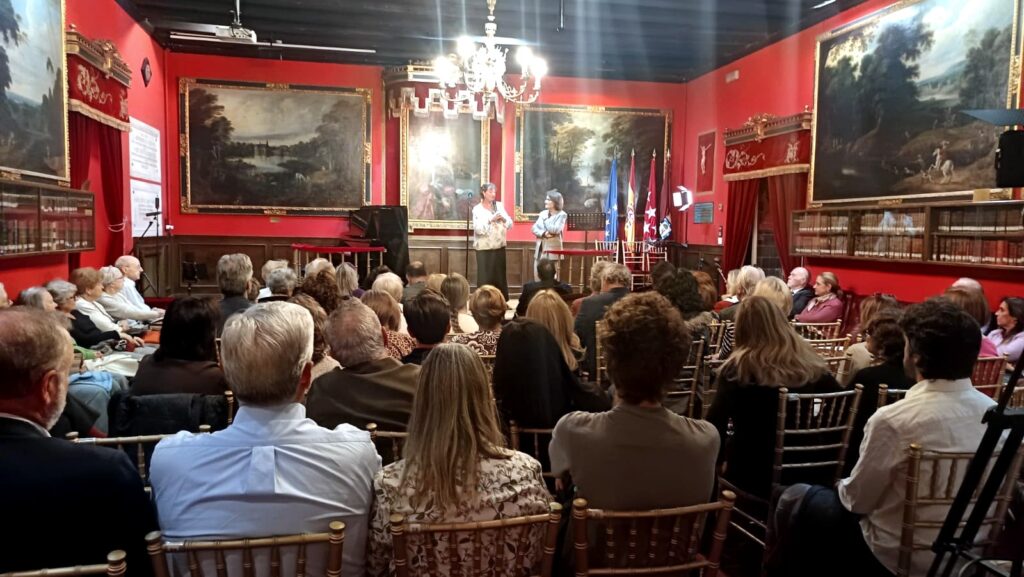 Imagen amplia del sal?n hist?rico de la Real Sociedad Econ?mica Matritense durante un acto cultural, con las ponentes en el escenario y numeroso p?blico ocupando la sala.