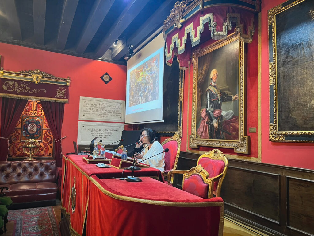 Una mujer habla por el micrófono desde una mesa roja mientras se proyecta una pintura en el salón histórico.