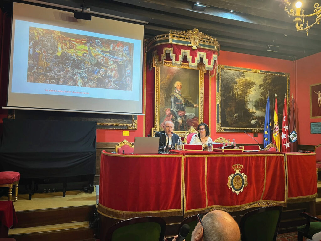 Dos participantes ante micrófonos en una mesa roja mientras se proyecta una pintura de Bernhard Heisig.