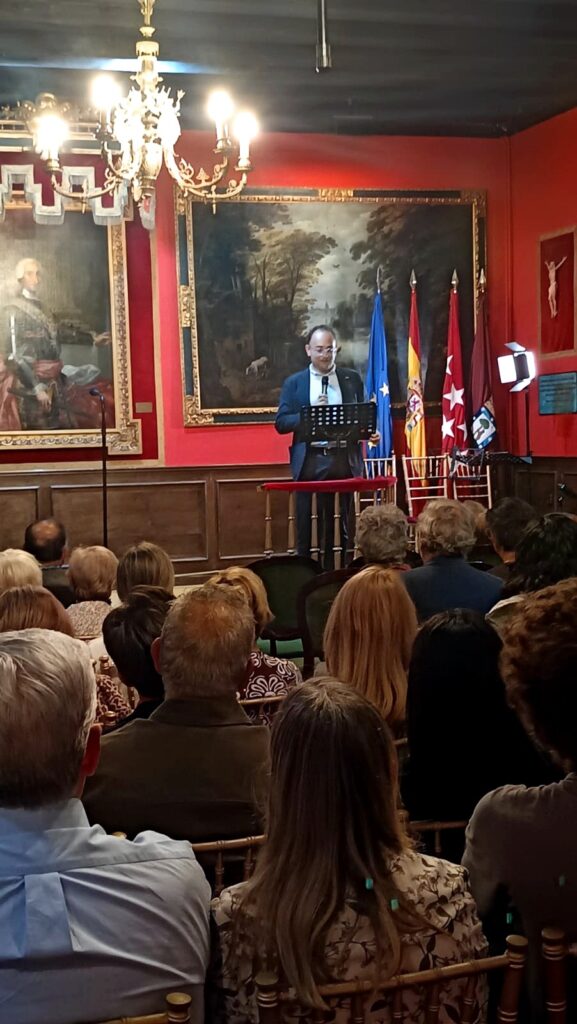 Un ponente interviene desde un atril en el sal?n hist?rico de la Real Sociedad Econ?mica Matritense, con el p?blico en primer plano y las banderas institucionales al fondo.