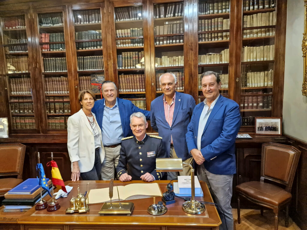 En una biblioteca revestida con altas vitrinas de madera repletas de volúmenes encuadernados, cuatro personas posan de pie alrededor de una persona sentada con uniforme azul oscuro