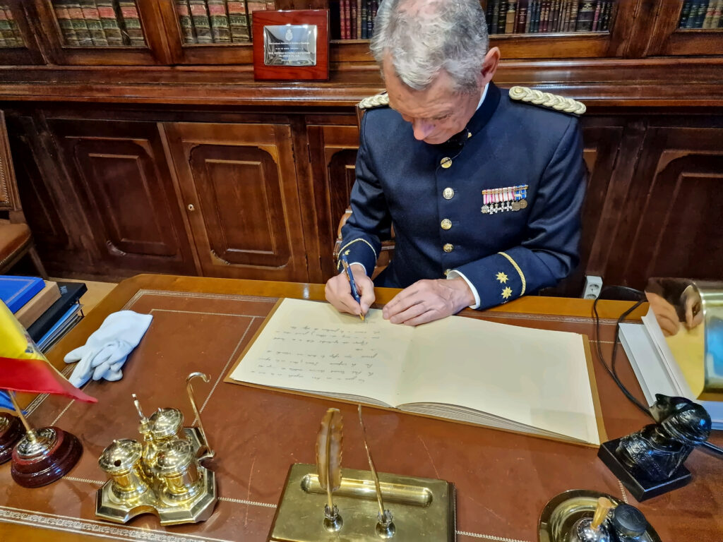 Plano ligeramente elevado de una persona de cabello gris, vestida con uniforme azul oscuro adornado con galones y medallas, mientras escribe con bolígrafo sobre la página izquierda