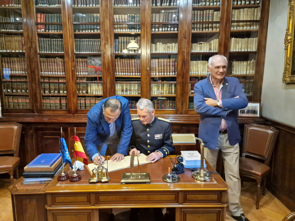 Fotografía horizontal tomada frente a un escritorio de madera. A la izquierda, un hombre con chaqueta azul se inclina para escribir en un libro abierto. En el centro, una persona c