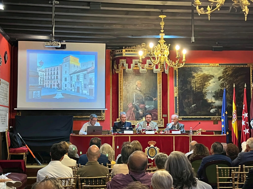 Panorámica frontal de un salón histórico de paredes rojas y techo de vigas oscuras. Cuatro participantes están sentados detrás de una mesa de terciopelo rojo con bordes dorados y e