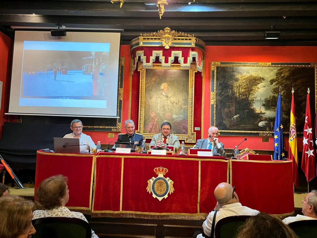 Plano frontal más cercano del estrado con cuatro personas sentadas ante una mesa institucional revestida de terciopelo rojo y adornos dorados. Sobre la mesa hay micrófonos, portáti
