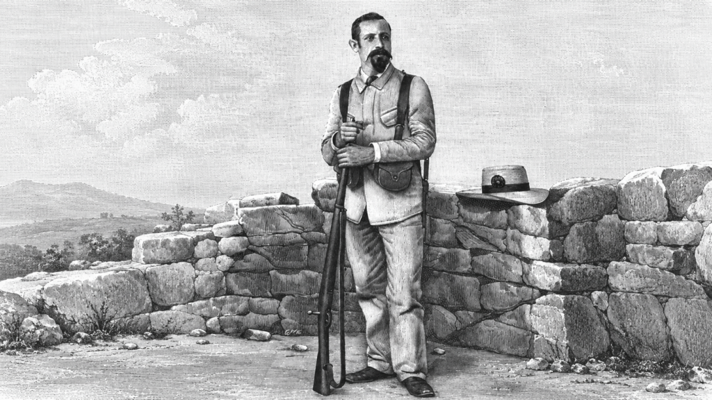 Hombre con barba y fusil largo junto a un muro de piedra.