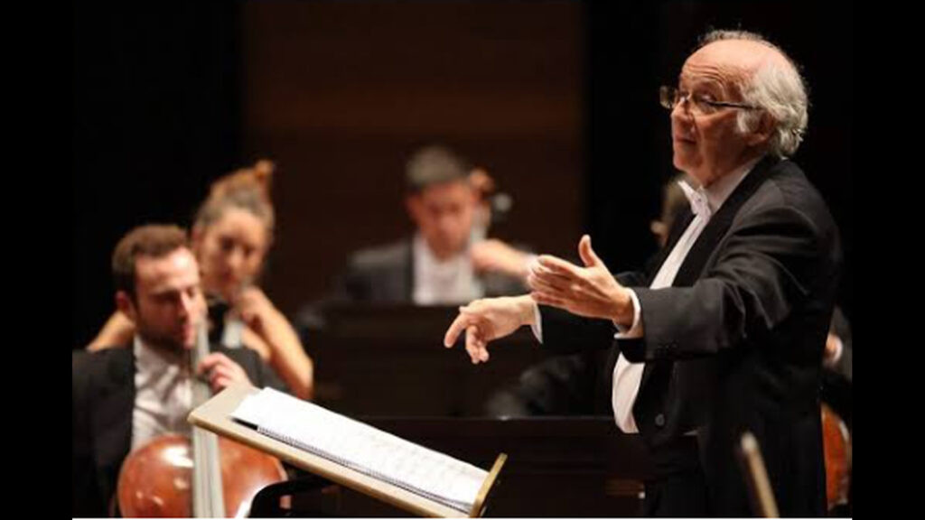 Un director de orquesta con gafas dirige con las manos ante varios músicos y una partitura abierta.